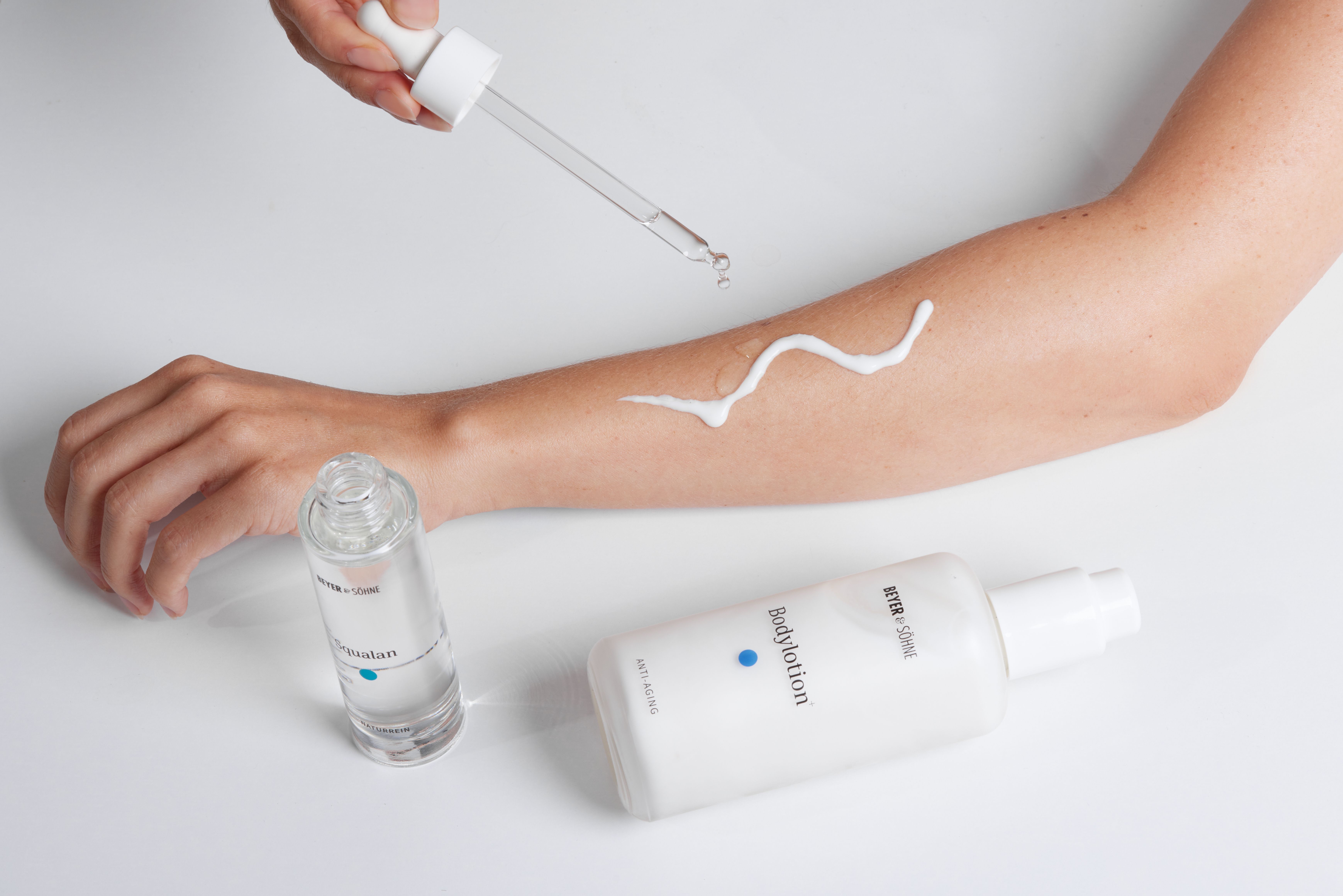 Ein Unterarm mit aufgetragener Bodylotion+ von Beyer & Söhne. Darüber wird mit einer Pipette Squalan aufgeträufelt. Die Flaschen der beiden Produkte liegen daneben.